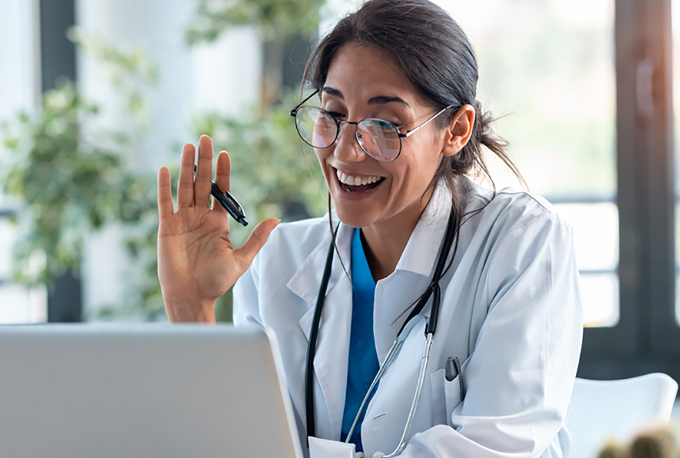 Female doctor waving and talking with colleagues through a video call with a laptop in the consultation.
